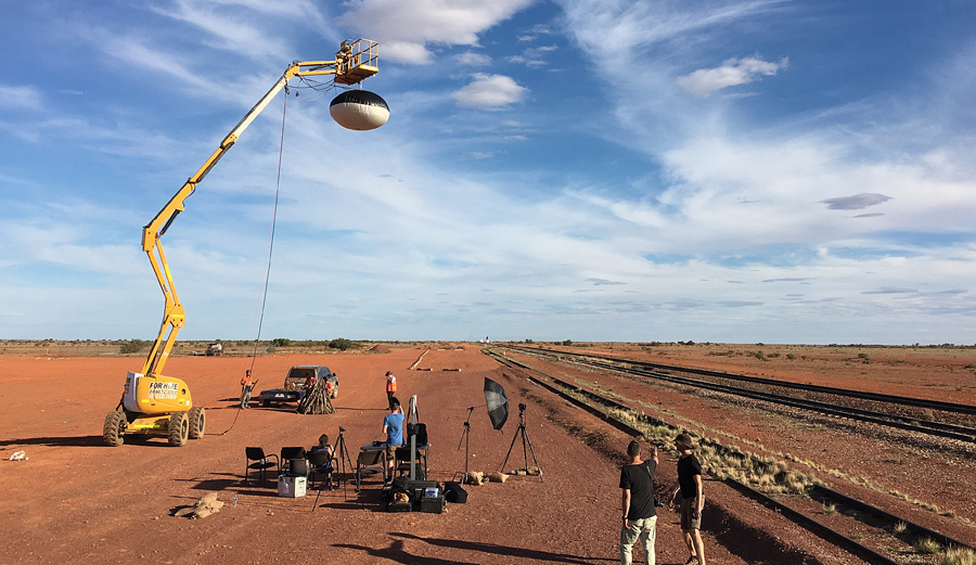 Title illustration: a film crew with equipment in the Australian outback. In front of the blue sky a blimp is hovering on a huge mobile crane over the red earth.